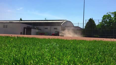 Low-angle-wide-shot-from-the-grass-field-of-a-tractor-grading-the-summer-time-ball-diamond,-kicking-up-dust
