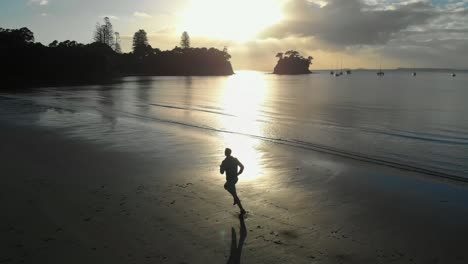 Cinematic-aerial-tracking-shot-of-silhouetted-young-man-running-on-a-beach-in-Auckland,-New-Zealand