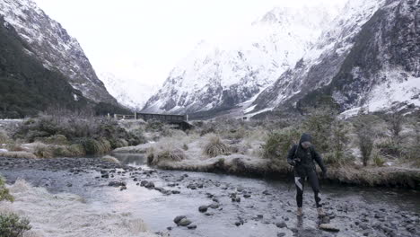 Slow-motion-shot-of-girl-in-hiking-gear-crossing-river-surrounded-by-snow-capped-mountains
