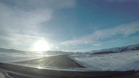 Perspectiva-Desde-El-Parabrisas-En-Un-Viaje-Por-Carretera-A-Través-De-Las-Nevadas-Montañas-Islandesas-Conduciendo-Hacia-La-Puesta-De-Sol