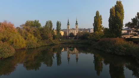 Establishing-shot-of-a-chatolic-church-in-a-park,-reflection-on-the-water-and-traffic-in-distance