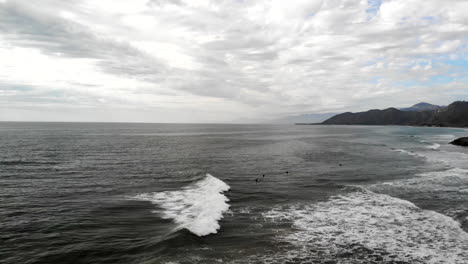 Aerial-drone-shot-on-a-cloudy-dark-epic-day-over-a-group-of-surfers-in-wetsuits-waiting-for-waves-to-surf-in-the-Pacific-Ocean-off-the-coast-of-Ventura,-California