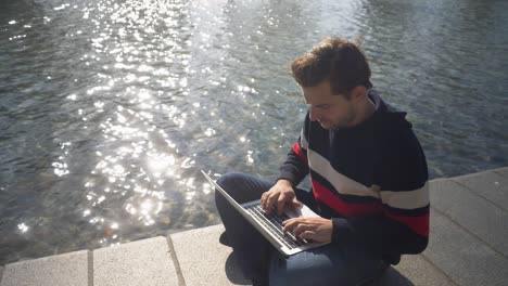 Nice-looking-young-man-sitting-on-a-bank-of-the-canal-in-Amsterdam-typing-on-the-notebook