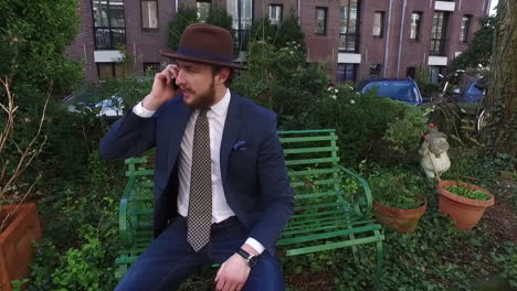 An-elegant-man-in-dandy-style-with-blue-suit-and-beard-is-talking-on-a-cellphone-on-a-green-bench-in-Amsterdam