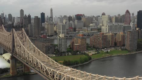 Rising-Drone-aerial-shot-looking-across-Roosevelt-Island-next-to-the-Queensboro-Bridge-at-the-East-River-towards-Manhattan,-NYC-in-the-daytime