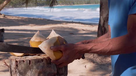 Close-up-shot-in-slow-motion-of-man-cutting-coconut-from-water,-with-turquoise-blue-beach-background-view-and-waves-hitting-the-sand-in-a-natural-environment