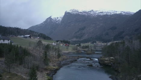 Flying-over-stream-in-Norwegian-wilderness-surrounded-by-mountains-and-houses,-Aerial-shot