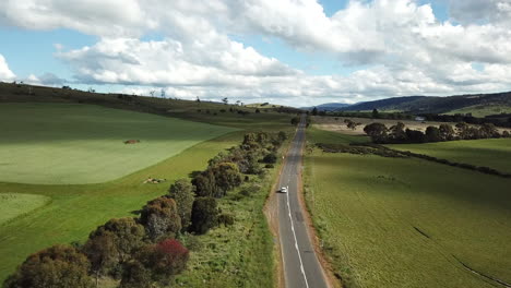 Drone-Shot-Of-Car-Driving-Into-Distance-Along-Country-Road-Lined-By-Trees-And-Fields
