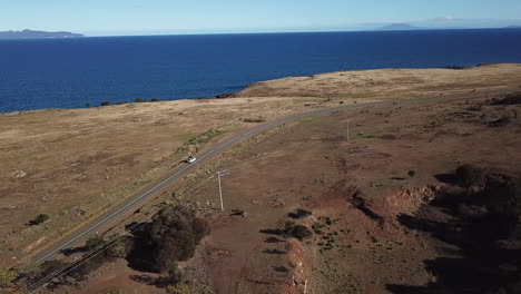Drone-Pan-Around-Lone-Car-Driving-Along-Coastal-Road-With-Trees-And-Blue-Ocean,-Tasmania-Australia