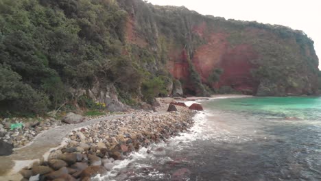 FPV-aerial-flight-alongside-stunning-red-cliffs-and-stone-path-next-to-beautiful-turquoise-ocean-water