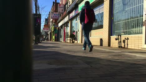 Low-angle-wide-shot-of-sidewalk-and-storefronts,-with-pedestrian-walking-by,-looking-westward-on-a-sunny-afternoon-on-College-Street-in-Toronto