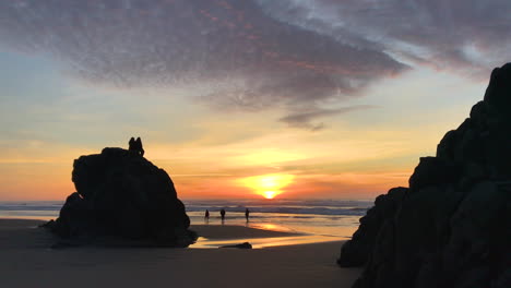 Leute,-Die-Auf-Felsen-Sitzen-Und-Bei-Sonnenuntergang-Am-Strand-An-Der-Küste-Von-Oregon-Spazieren-Gehen