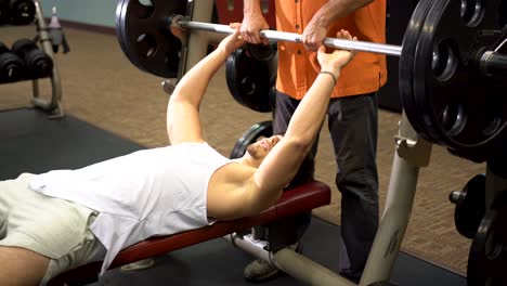 Teen-male-bodybuilder-lying-down-and-pressing-large-barbell-as-a-man-in-organge-spots-him