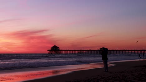 A-man-takes-a-picture-of-the-beach-during-a-gorgeous-red,-purple,-tangerine,-pink-and-blue-sunset-with-the-Huntington-Beach-Pier-in-the-background-at-Surf-City-USA-California