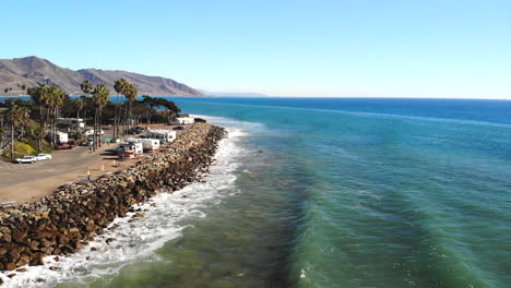 Aerial-drone-shot-rising-up-over-ocean-waves-crashing-on-the-rocky-coast-with-palm-trees-and-rv-campground-on-a-California-beach