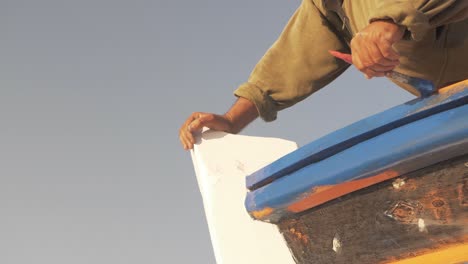Painter-touching-up-wooden-trim-of-traditional-Greek-carvel-build-fishing-boat-blue-paint-bow-slow-motion-low-angle