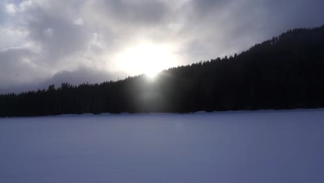 Schnee-Weht-Bei-Sonnenuntergang-über-Den-Zugefrorenen-See-Mit-Bäumen-Im-Hintergrund