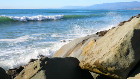 A-slow-motion-sliding-shot-of-large-blue-ocean-waves-crashing-against-the-rocks-on-a-California-beach-SLIDE-LEFT