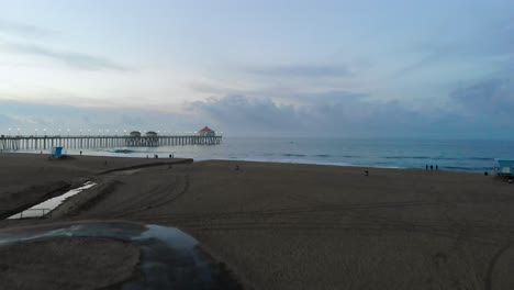 Mit-Dem-Pier-In-Der-Surf-City-Usa-Kalifornien-Bei-Sonnenaufgang-über-Den-Sand-Zum-Meer-Gleiten,-Während-Die-Menschen-Ihren-Urlaub-Genießen-Und-Surfer-Wellen-Fangen
