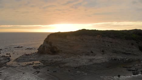 Aerial-shot-flying-backwards-while-looking-at-rugged-cliffs-on-Bass-coast-Victoria-Australia-with-the-sunset-on-the-horizon