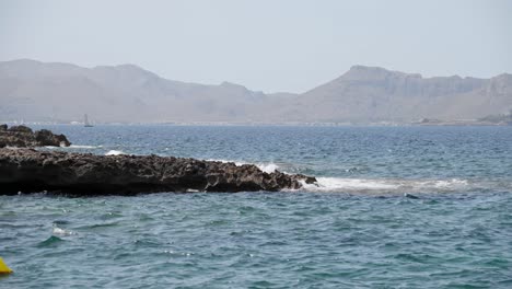 Felsenstrand,-Umgeben-Von-Bergen-Mit-Einigen-Yachten-Im-Hintergrund-Auf-Der-Insel-Mallorca