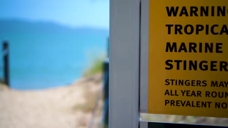 Close-up-shot-of-marine-stinger-warning-sign-before-camera-slides-to-the-left-revealing-beach-access-track-and-calm-blue-ocean