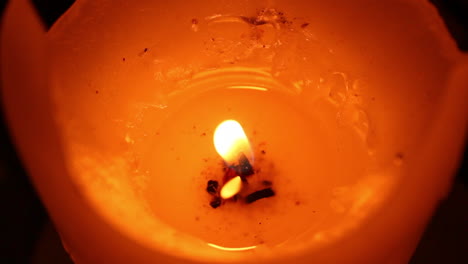 Angled-close-view-of-an-orange-and-red-candle-with-a-litted-shaky-flame-and-melted-wax-on-dark-background
