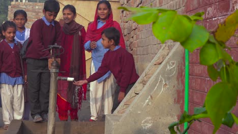 A-group-of-Pakistani-rural-area-school-children-using-a-hand-pump,-In-winter`s-season,-wearing-sweaters-and-jackets,-Fun-and-smiling,-Red-bricks-wall-and-an-old-gate-of-the-school