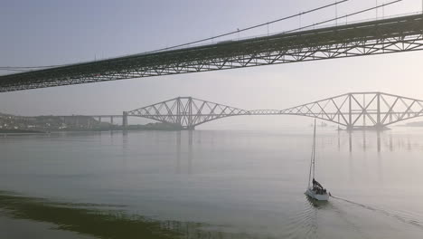 Aerial-footage-of-a-sailing-boat-travelling-beneath-the-old-Forth-Road-Bridge-at-South-Queensferry-on-a-sunny-day-in-West-Lothian,-Scotland