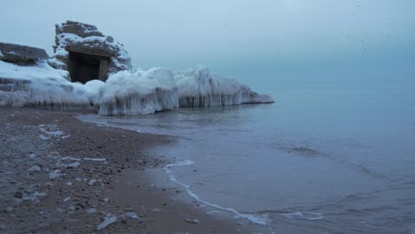Small-waves-breaking-against-the-ruins-of-Karosta-Northern-Forts-fortification-on-the-shore-of-Baltic-sea-on-a-cloudy-winter-day,-covered-with-ice,-snow-and-icicles,-wide-shot
