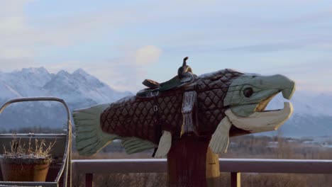 Sunrise-in-Alaska,-pan-around-wooden-trout-statue-with-saddle-revealing-beautiful-distance-mountains-of-Anchorage-4k