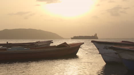 Alte-Fischerboote-In-Port-Royal-Bei-Sonnenuntergang-Mit-Containerschiff-Im-Hintergrund