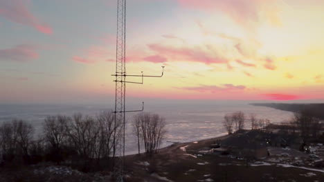 A-wind-meter-blowing-on-a-tower-with-a-beautiful-orange-and-pink-sunset-and-frozen-winter-lake-in-the-background