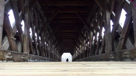 Low-angle-on-lone-figure-exiting-walkway-at-other-end-of-old,-wooden,-covered-river-bridge