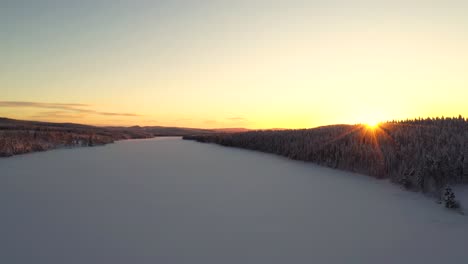 Aerial-view-of-sunset-over-snowy-frozen-lake-in-deep-forest-with-hills-and-mountains
