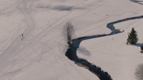 Drone-Orbit-shot-of-a-man-cross-country-skiing-with-his-dog-in-a-snowy-landscape-with-a-river
