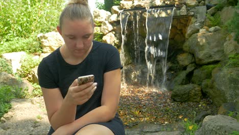 Beautiful-Woman-In-Nature-Uses-Her-Mobile-Device-While-Sitting-On-A-Park-Bench-Next-To-A-Waterfall