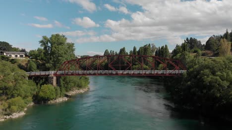 SLOWMO---Aerial---Red-bridge-over-beautiful-blue-river-in-Clyde,-Central-Otago,-New-Zealand-with-cyclists-biking-over