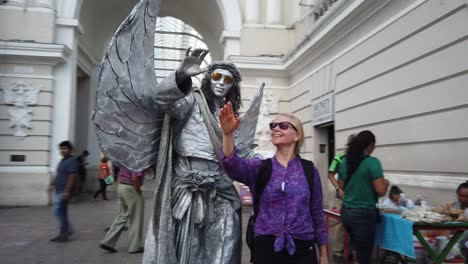 An-angel-mime-statue-indicating-taking-a-photo-with-him,-a-mature-woman-gets-next-to-him-and-mimics-his-movements