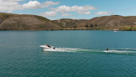 Waterskiing-behind-boat-on-Lake-Dunstan-near-Clyde-dam,-Central-Otago,-New-Zealand-with-mountains-and-clouds-in-background