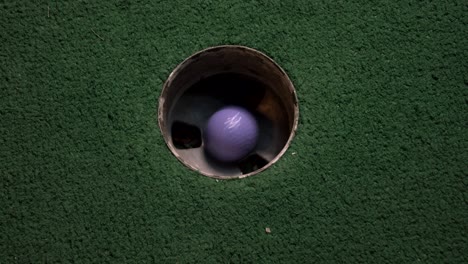A-close-up-birds-eye-view-of-a-purple-mini-golf-ball-falls-into-the-golf-hole-and-bounces-in-the-pocket-on-a-course