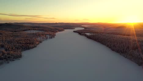 Aerial-view-of-sunset-over-snowy-frozen-lake-in-deep-forest-with-hills-and-mountains