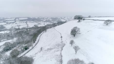 Aerial-forward-tracking-across-vast-English-farmland-covered-in-snow,-with-a-hillside-common-in-the-foreground-with-a-valley-in-the-background