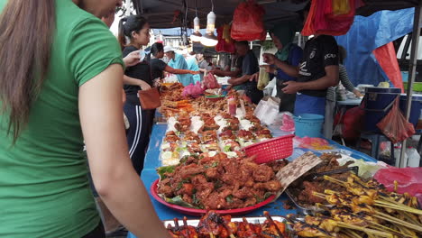 Many-people-standing-and-buying-food-from-the-seller-with-loads-of-different-kind-of-meat-on-the-table-with-hanging-plastic-bags-and-light-bulbs