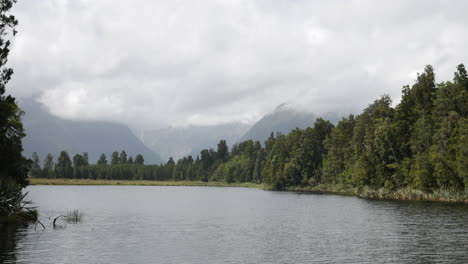 Panoramic-shot-of-a-river-flowing,-surrounded-by-a-forest,-in-New-Zealand,-with-mountains-covered-by-clouds-in-the-background,-on-a-cloudy-day