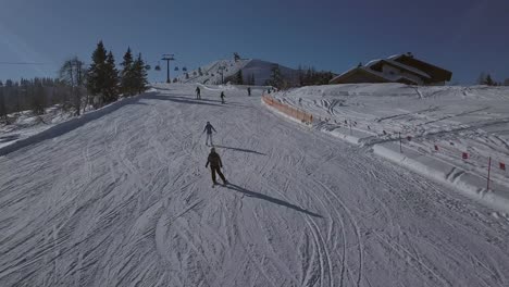 AERIAL-dolly-shot-of-a-ski-slope-in-a-ski-resort-on-a-sunny-day-with-people-on-the-slope-and-they-are-following-the-camera-down-the-slope-with-more-people-and-beautiful-mountains-in-the-background