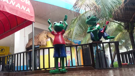 Two-Green-Frog-Statues-Wearing-Clothes-Outside-Of-Senior-Frogs-In-Mexico-During-The-Rain