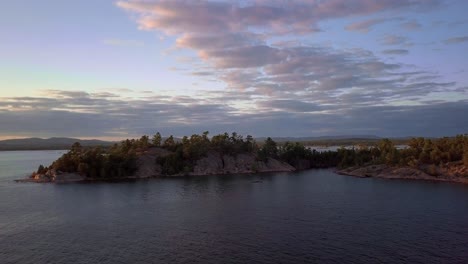 Fly-to-a-Rocky-Pine-Tree-Island,-Blue-Sky-and-Clouds-at-Sunset,-Drone-Aerial-Wide-Dolly-In
