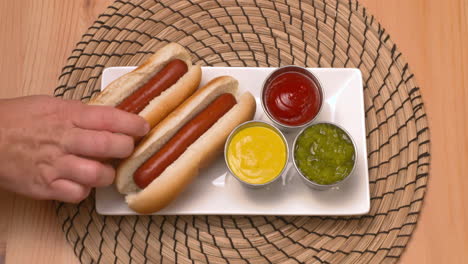 Top-down-view-of-a-hand-placing-hot-dogs-on-a-plate-beside-ketchup,-mustard,-and-relish