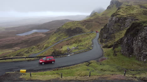 Panning-shot-from-a-vista-of-Rocky-cliffs,-meadows-and-lakes-with-curvy-road-in-the-foreground-with-passing-cars-on-a-rainy-and-cloudy-day-in-Quiraing,-Scotland,-Isle-of-Skye